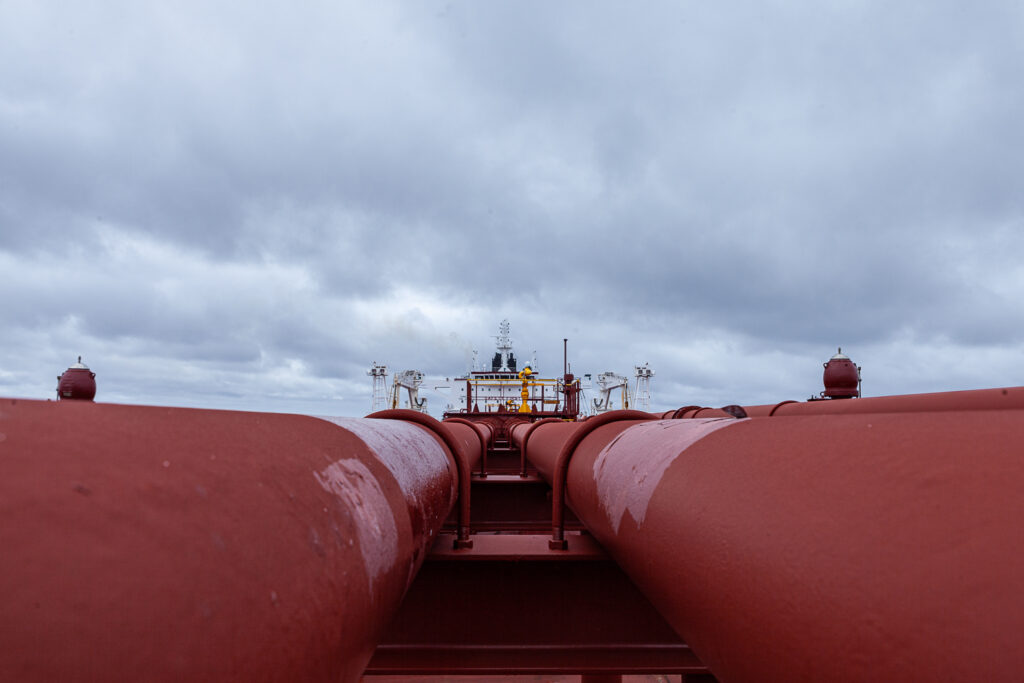 View onboard a tanker vessel, standing between large red pipes with the bridge in the distance.