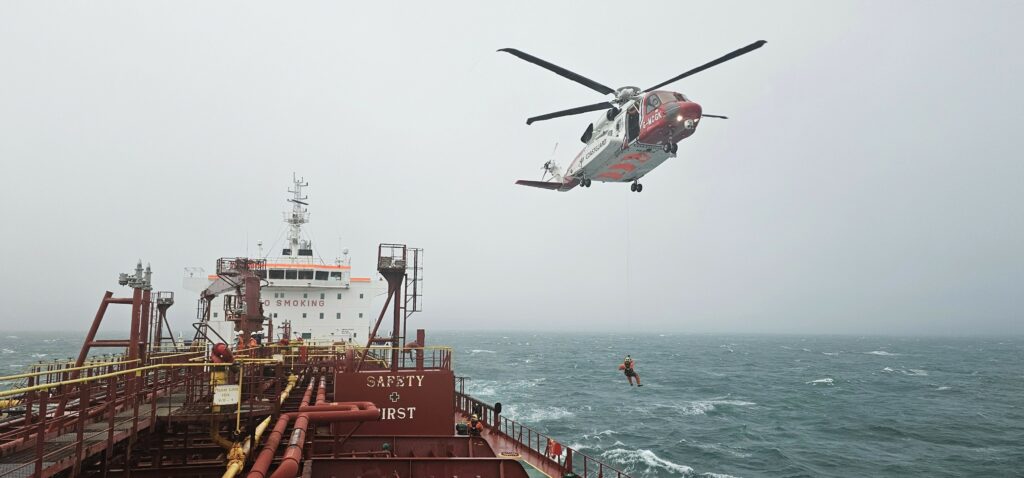 A helicopter flying above a tanker vessel with the words 'Safety First'. A person is hanging from the helicopter on a safety rope.